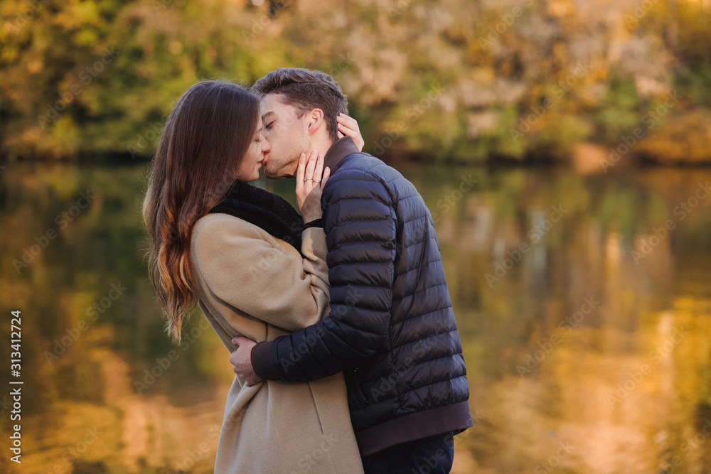 Couple in love standing in autumn park with yellow fallen leaves. Man and woman enjoying a day together. Boy kissing a girl in lips Couple in love standing in autumn park with yellow fallen leaves. Man and woman enjoying a day together. Boy kissing a girl in lips
