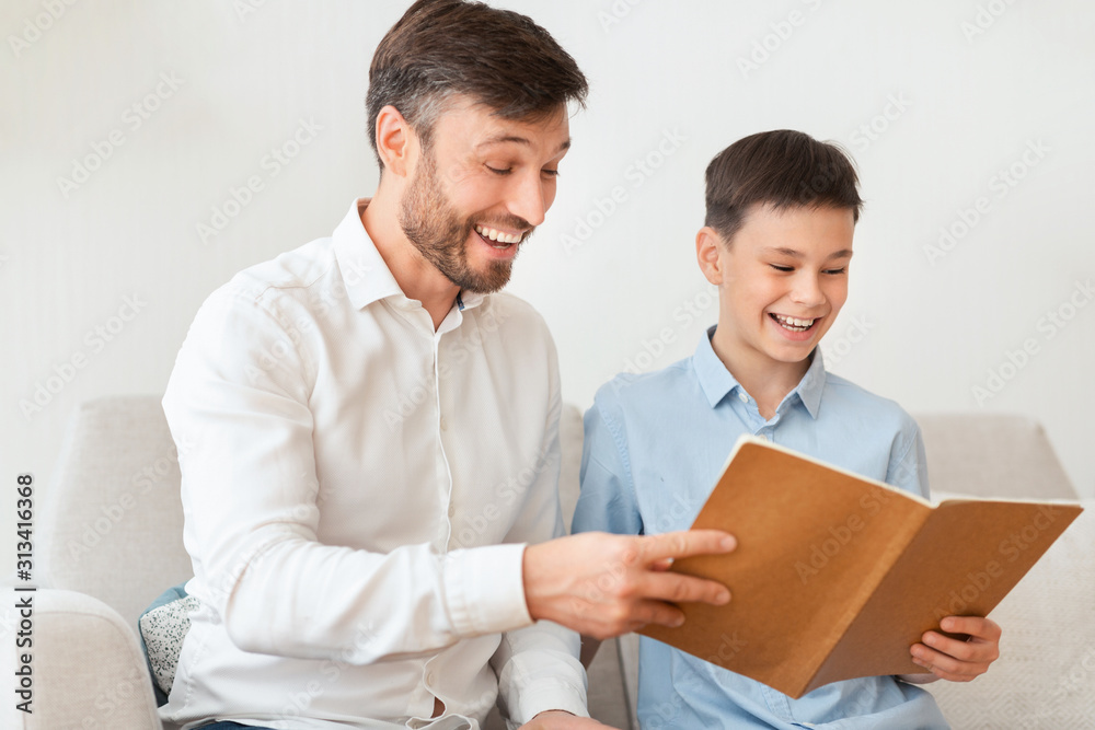 Father And Son Reading Book Sitting On Couch At Home Father And Son Reading Book Sitting On Couch At Home