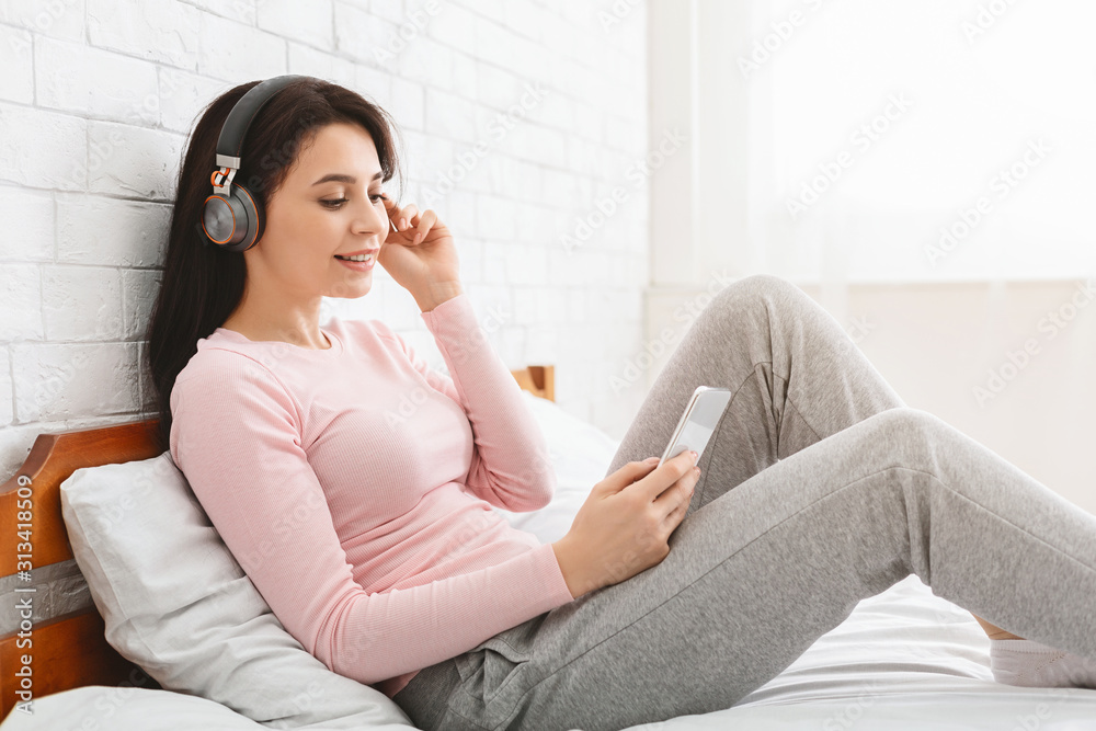 Cheerful girl in headphones typing on cellphone, lying in bed Cheerful girl in headphones typing on cellphone, lying in bed