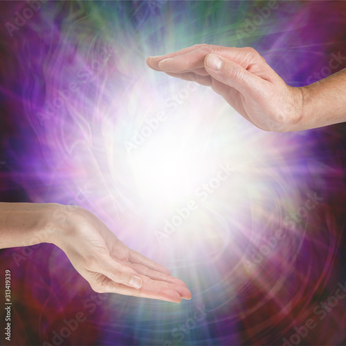 Together our healing energies combine to create a powerful influence - male hand opposite female hand with a white light orb in between against a multicoloured flowing energy field background with cop photo