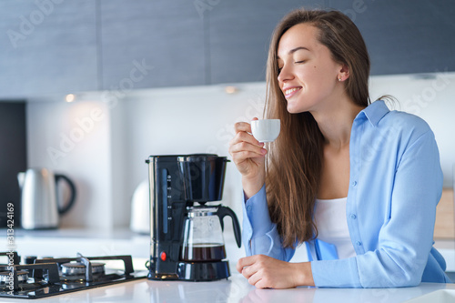 Leinwand Poster Happy attractive woman enjoying of fresh coffee aroma after brewing coffee using coffee maker in the kitchen at home