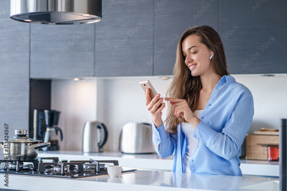 Young happy woman using smartphone and wireless headphones for listening music and making video calls in the kitchen at home. Modern mobile people