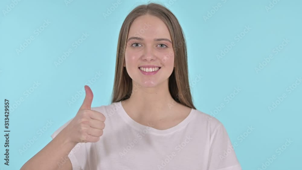 Thumbs Up by Cheerful Beautiful Young Woman, Blue Background Thumbs Up by Cheerful Beautiful Young Woman, Blue Background