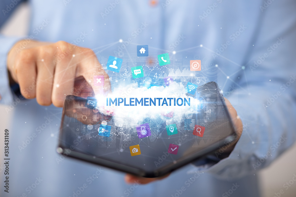 Businessman holding a foldable smartphone with IMPLEMENTATION inscription, technology concept Businessman holding a foldable smartphone with IMPLEMENTATION inscription, technology concept