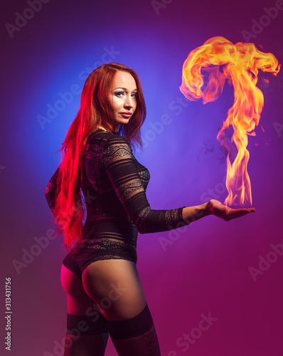 Sexy young woman in black transparent erotic costume and stockings lights a flame on her hand in the studio