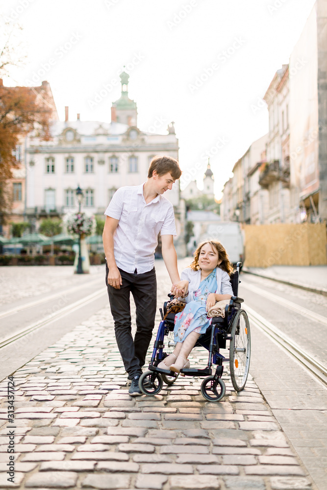 Young Couple In Wheelchair Strolling In old City. Lovely couple, woman in wheelchair and her husband, walking outdoors in the city