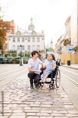 Young smiling woman in wheelchair and joyful man walking outdoors in old city center in summer morning