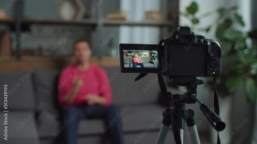 Close-up of professional video camera on tripod filming positive hearing impaired male blogger in domestic room. Deaf-mute web influencer recording video for vlog using sign language at home.
