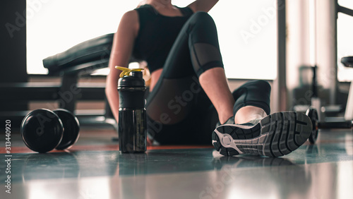 Fototapeta Naklejka Na Ścianę i Meble -  fitness ,workout, gym exercise ,lifestyle  and healthy concept. Side view of a woman sitting to relax after a workout with a whey protein and dumbbell and an apple placed beside in the gym at sunset.