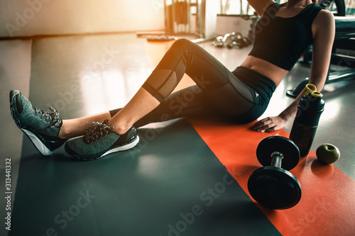 fitness ,workout, gym exercise ,lifestyle  and healthy concept. Side view of a woman sitting to relax after a workout with a whey protein and dumbbell and an apple placed beside in the gym at sunset. photo