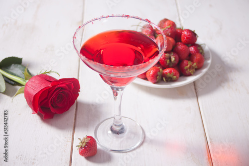 Red exotic alcoholic cocktail in clear glass, plate with fresh strawberries and red rose on the wooden white table for romantic dinner.