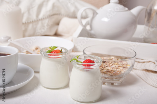 Two portions natural homemade yogurt in a glass jar with fresh strawberry and muesli, teapot, cup of coffee nearby. Breakfast concept.