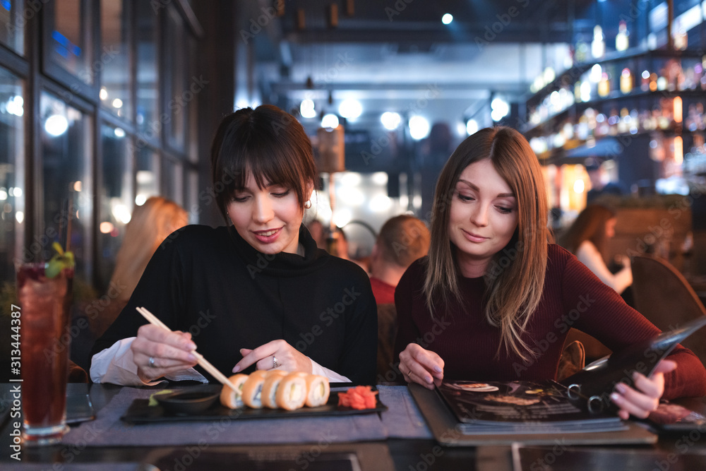 Fototapeta premium Girls eating a dinner at restaurant. Brunette is eating sushi and her girlfriend is ordering food from a menu.