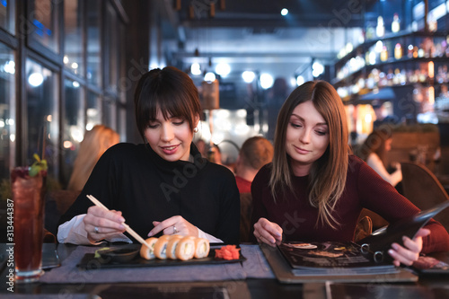 Girls eating a dinner at restaurant. Brunette is eating sushi and her girlfriend is ordering food from a menu.
