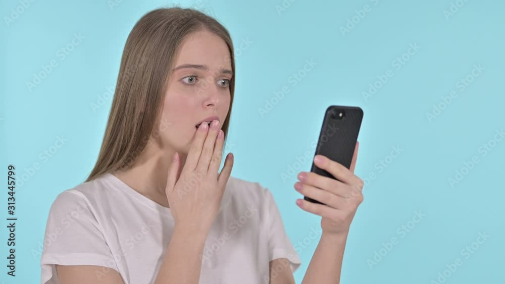 Young Woman Upset by Loss on Smartphone, Blue Background Young Woman Upset by Loss on Smartphone, Blue Background