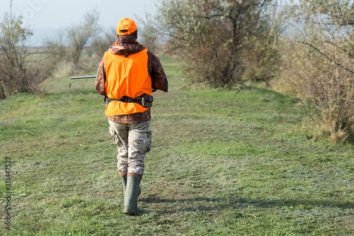 A man with a gun in his hands and an orange vest on a pheasant hunt in a wooded area in cloudy weather. Hunter with dogs in search of game. © Mountains Hunter