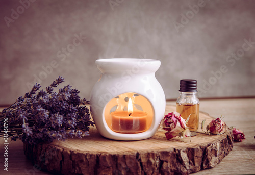 Vintage style picture of white ceramic candle aroma oil lamp with essential oil bottle and dry flower petals on natural pine wood disc, dry background with copy space. photo