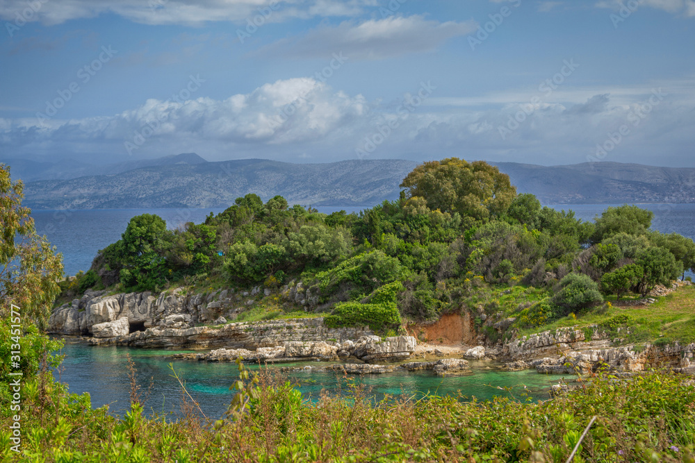 Beautiful landscape – sea lagoon with turquoise water, cliffs and rocks, covered with trees green grass and bushes, blue sky and mountains on the horizon. Corfu Island, Greece. 