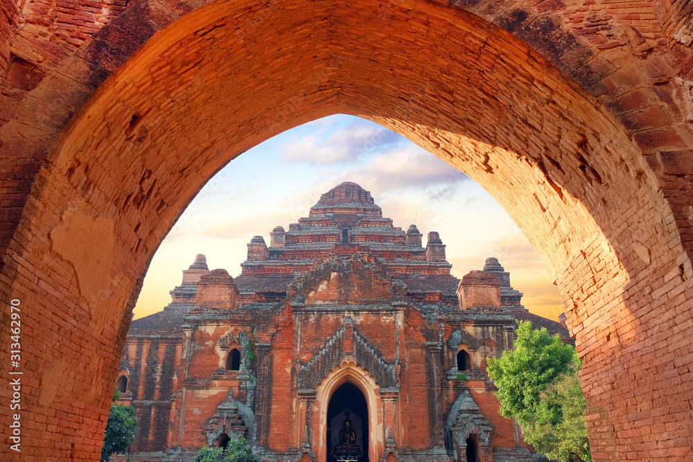 Entry gate in an old temple surrounded by green vegetation in old Bagan, Myanmar, under a colorful sunrise sky.
