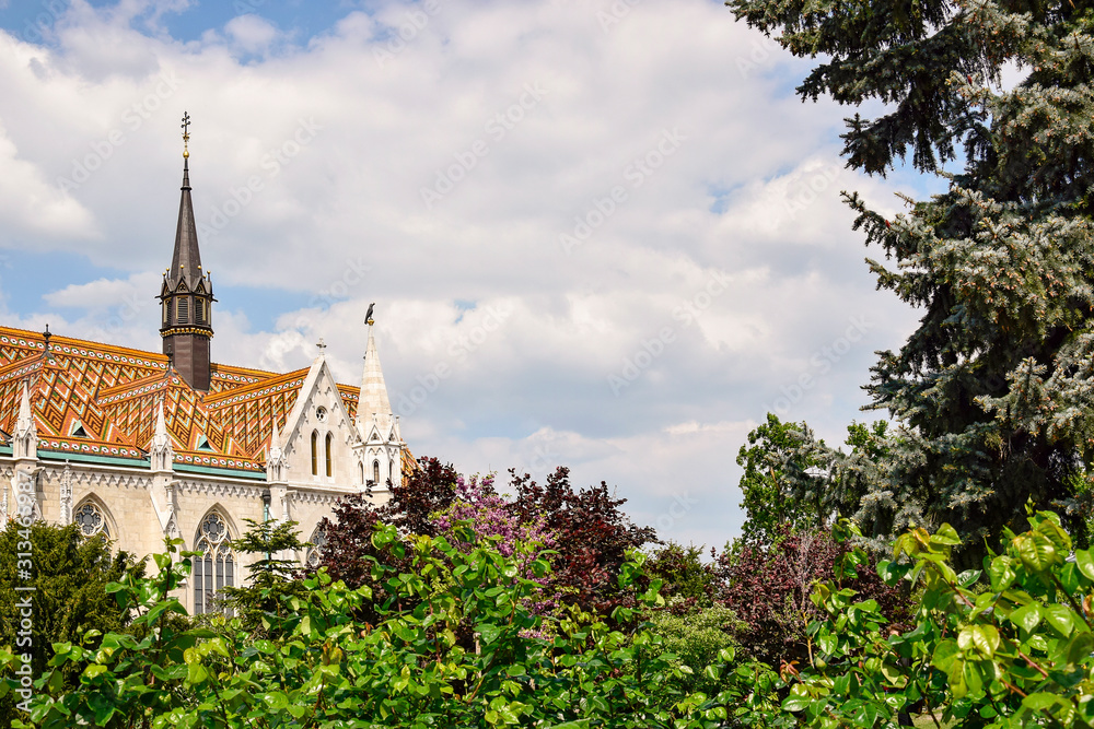 View of ornately colored tile roof of St. Matthias Church through the lush greens of trees on cloudy sky background. Fisherman's Bastion in Budapest.