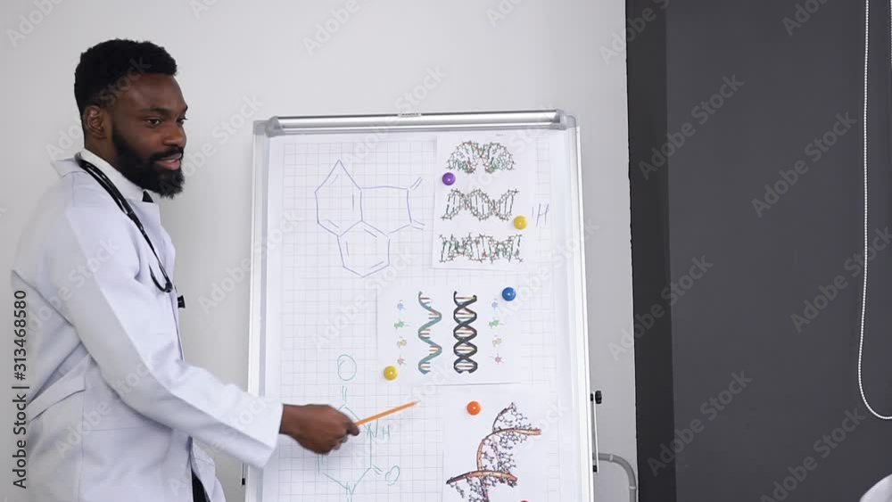 Attractive young bearded dark-skinned doctor lecturing and explaining the chemical compounds on the poster in the hospital cabinet Attractive young bearded dark-skinned doctor lecturing and explaining the chemical compounds on the poster in the hospital cabinet