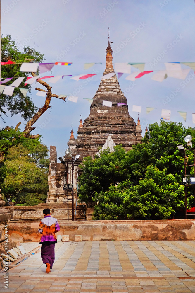 Colorful praying flags and person walking in front of an ancient Buddhist temple surrounded by vegetation in Bagan, Myanmar.