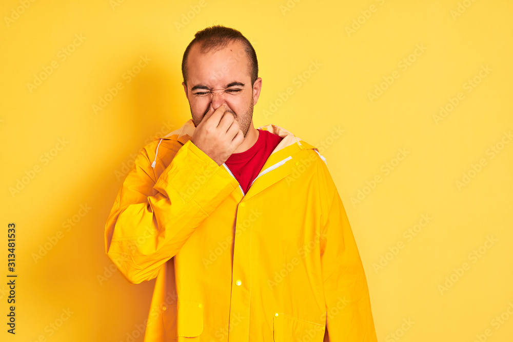 Young man wearing rain coat standing over isolated yellow background smelling something stinky and disgusting, intolerable smell, holding breath with fingers on nose. Bad smells concept. Young man wearing rain coat standing over isolated yellow background smelling something stinky and disgusting, intolerable smell, holding breath with fingers on nose. Bad smells concept.
