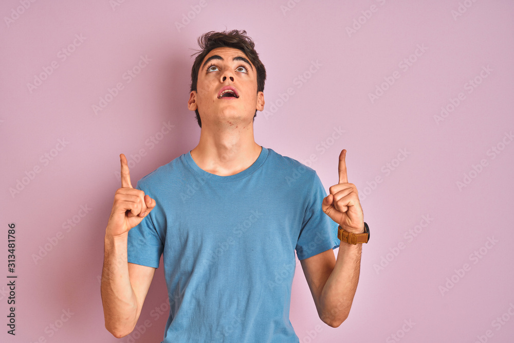 Teenager boy wearing casual t-shirt standing over blue isolated background amazed and surprised looking up and pointing with fingers and raised arms. Teenager boy wearing casual t-shirt standing over blue isolated background amazed and surprised looking up and pointing with fingers and raised arms.
