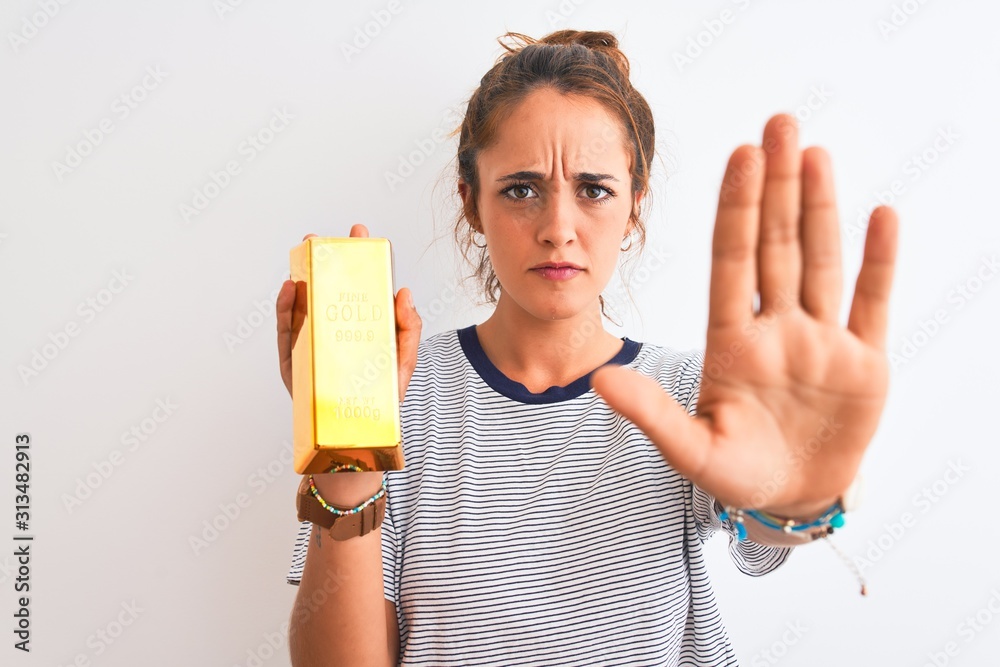 Young beautiful redhead woman holding gold ingot over isolated background with open hand doing stop sign with serious and confident expression, defense gesture