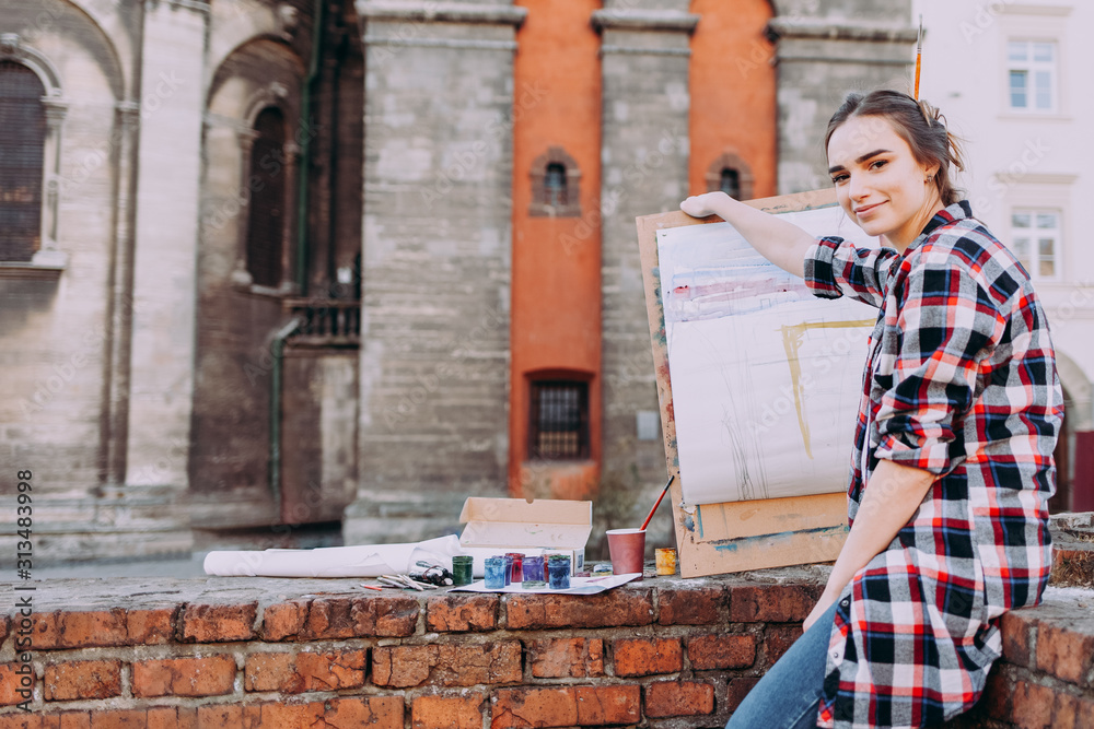 Woman artist paints a picture while sitting on a brick wall on a background of old architecture. Girl spends leisure drawing pictures on a city street. Smiling female artist