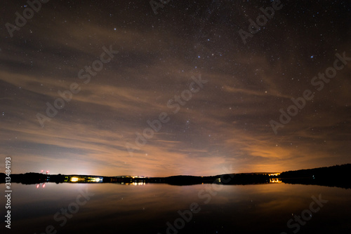 starry sky with some clouds over a lake and lights on the horizon