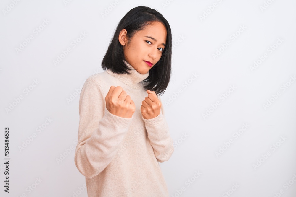 Young chinese woman wearing turtleneck sweater standing over isolated white background Ready to fight with fist defense gesture, angry and upset face, afraid of problem Young chinese woman wearing turtleneck sweater standing over isolated white background Ready to fight with fist defense gesture, angry and upset face, afraid of problem