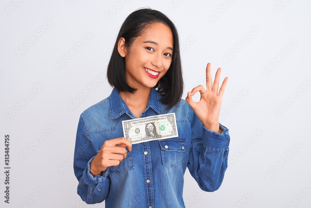 Young beautiful chinese woman holding one dollar standing over isolated white background doing ok sign with fingers, excellent symbol Young beautiful chinese woman holding one dollar standing over isolated white background doing ok sign with fingers, excellent symbol