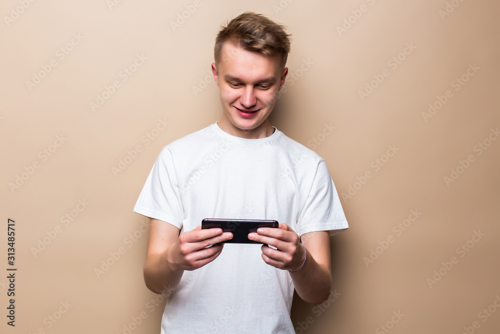 Portrait a happy young man playing games on mobile phone standing isolated over beige background