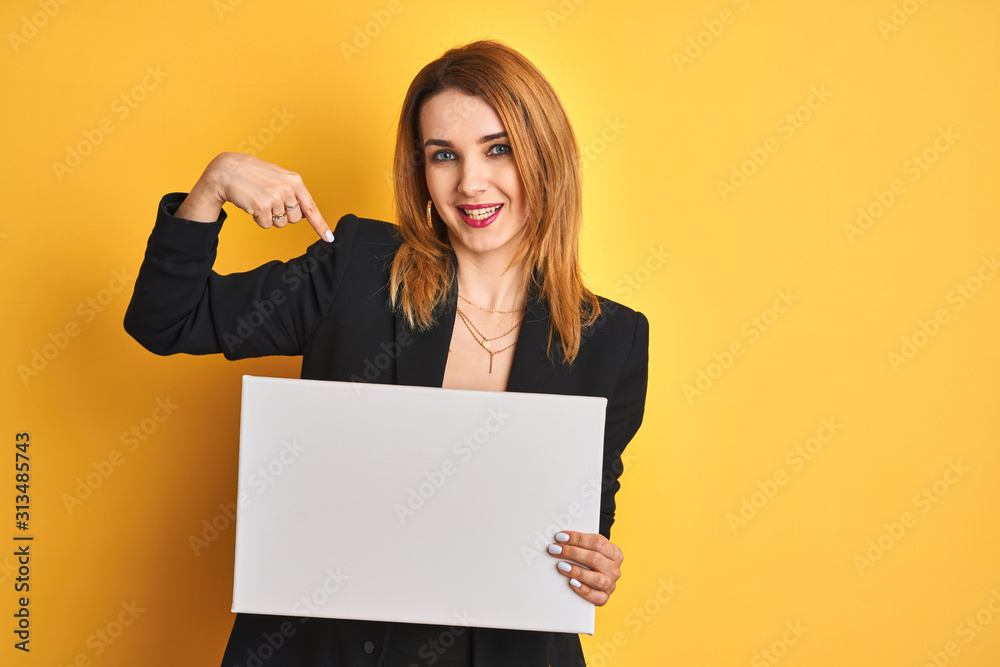 Naklejka premium Redhead business caucasian woman holding banner over yellow isolated background with surprise face pointing finger to himself