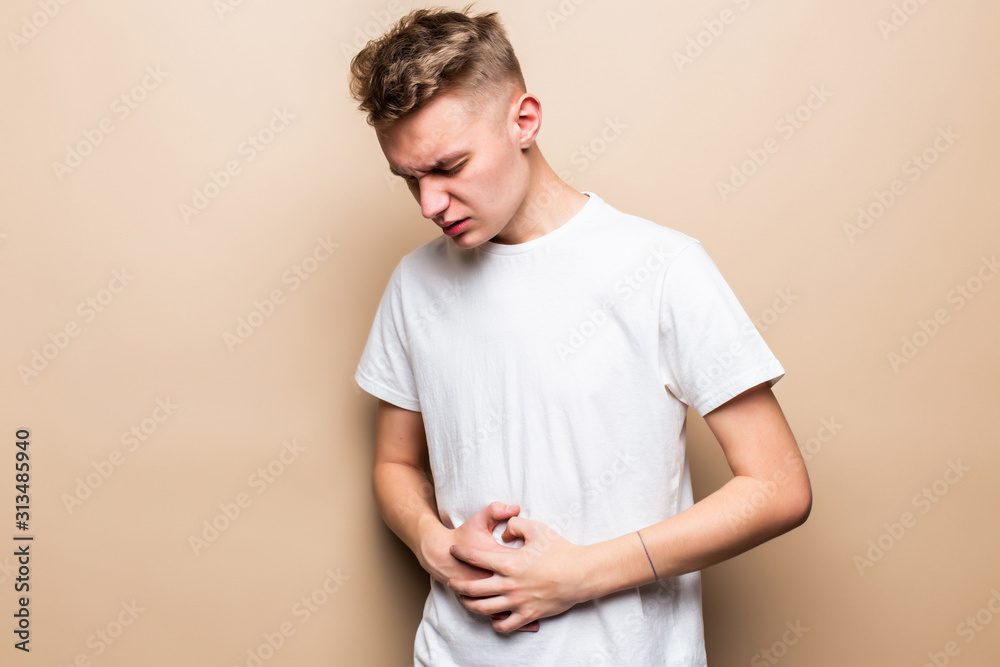 Young teen man suffers from pain in stomack both hands isolated on beige background. Young teen man suffers from pain in stomack both hands isolated on beige background.