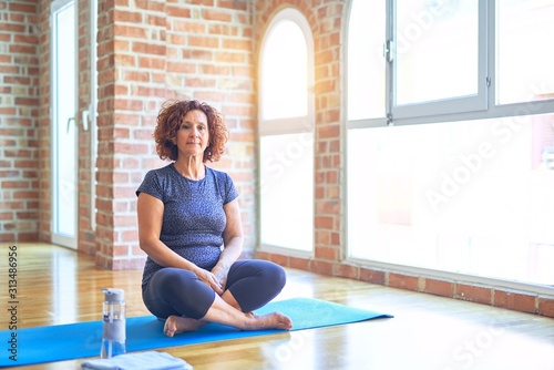Middle age beautiful sportswoman wearing sportswear sitting on mat practicing yoga at home Relaxed with serious expression on face. Simple and natural looking at the camera.