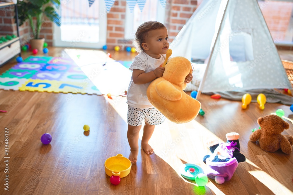 Adorable toddler holding duck doll standing around lots of toys at kindergarten - obrazy, fototapety, plakaty 