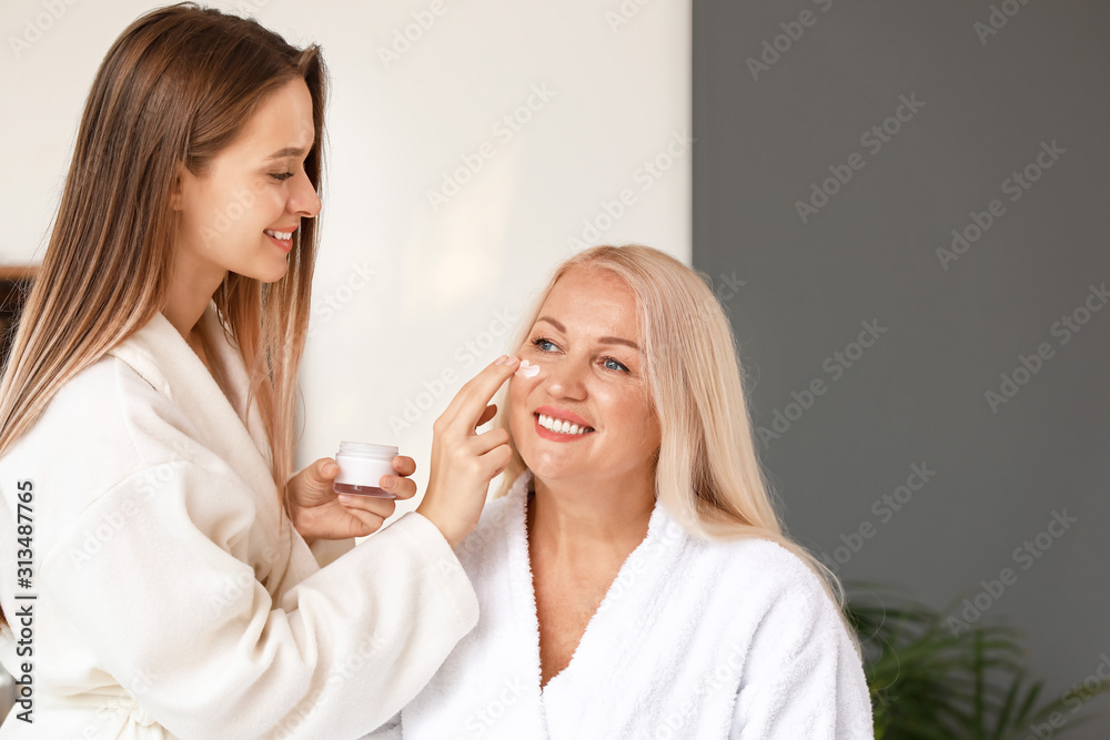 Adult daughter applying cream onto face of her mature mother in bathroom Adult daughter applying cream onto face of her mature mother in bathroom