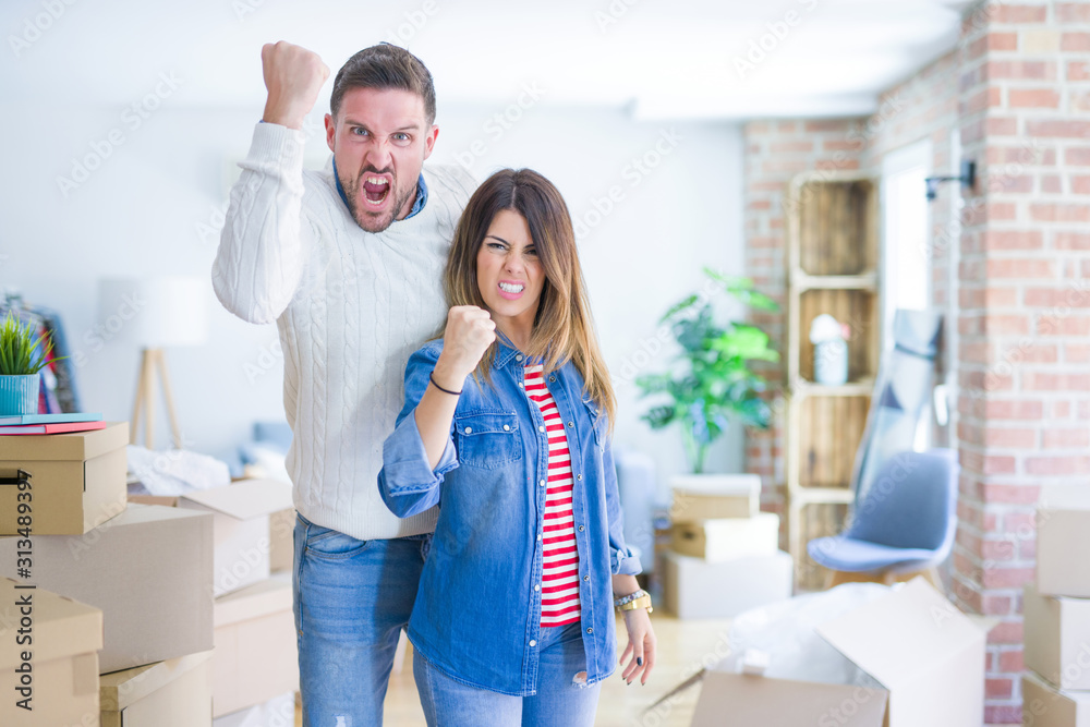 Young beautiful couple standing at new home around cardboard boxes angry and mad raising fist frustrated and furious while shouting with anger. Rage and aggressive concept. Young beautiful couple standing at new home around cardboard boxes angry and mad raising fist frustrated and furious while shouting with anger. Rage and aggressive concept.
