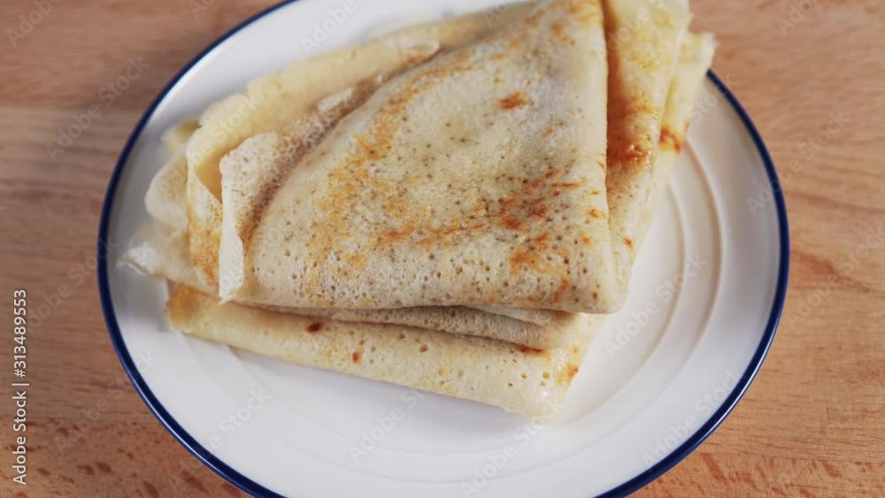 A hand picks up a freshly baked, lush, porous pancake from a heap on a white plate with a blue border. On a wooden table A hand picks up a freshly baked, lush, porous pancake from a heap on a white plate with a blue border. On a wooden table