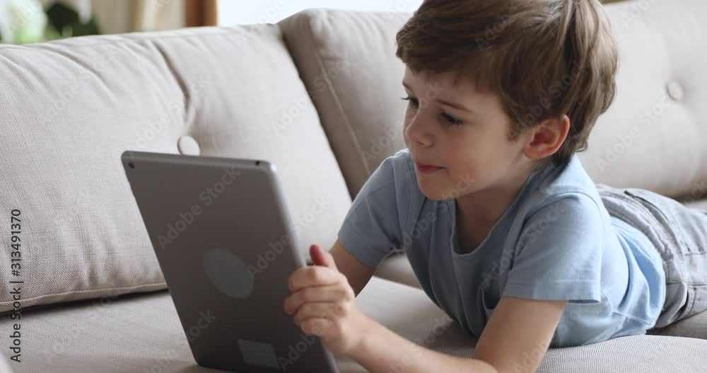 Happy cute preschool boy using tablet lying on sofa alone