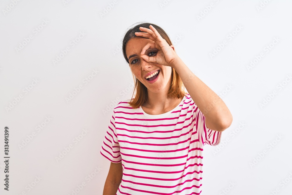 Beautiful redhead woman wearing casual striped pink t-shirt over isolated background doing ok gesture with hand smiling, eye looking through fingers with happy face. Beautiful redhead woman wearing casual striped pink t-shirt over isolated background doing ok gesture with hand smiling, eye looking through fingers with happy face.