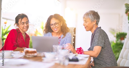 Meeting of middle age women having lunch and drinking coffee. Mature friends smiling happy using laptop at home on a sunny day