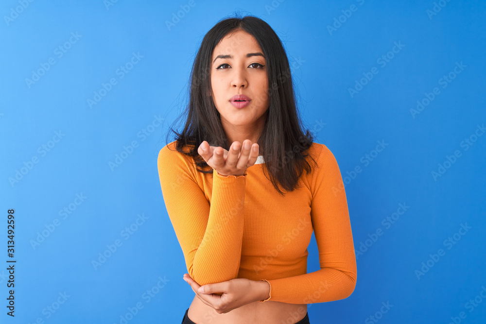 Young beautiful chinese woman wearing orange t-shirt standing over isolated blue background looking at the camera blowing a kiss with hand on air being lovely and sexy. Love expression. Young beautiful chinese woman wearing orange t-shirt standing over isolated blue background looking at the camera blowing a kiss with hand on air being lovely and sexy. Love expression.