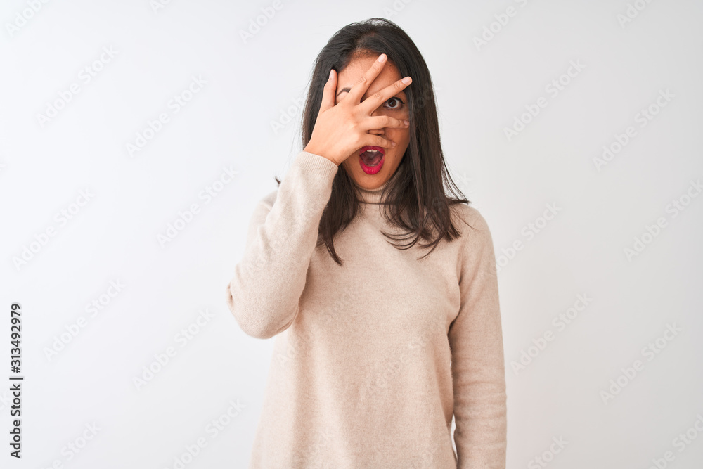 Beautiful chinese woman wearing turtleneck sweater standing over isolated white background peeking in shock covering face and eyes with hand, looking through fingers with embarrassed expression.