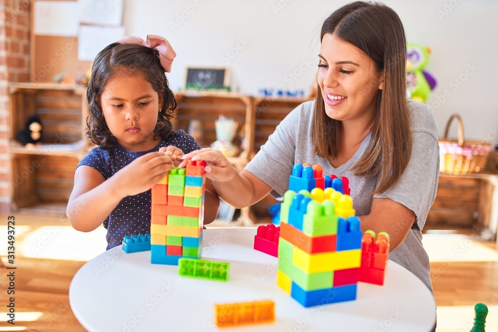 Beautiful teacher and toddler girl playing with construction blocks bulding tower at kindergarten - obrazy, fototapety, plakaty 