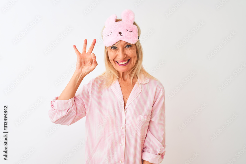 Middle age woman wearing sleep mask and pajama over isolated white background smiling positive doing ok sign with hand and fingers. Successful expression. Middle age woman wearing sleep mask and pajama over isolated white background smiling positive doing ok sign with hand and fingers. Successful expression.