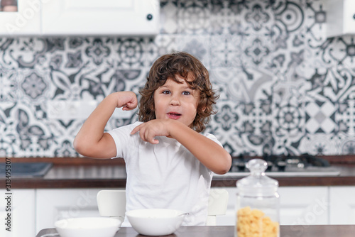 Happy little boy showing biceps muscles while eating healthy breakfast of milk and cornflakes at the stylish kitchen in the morning.