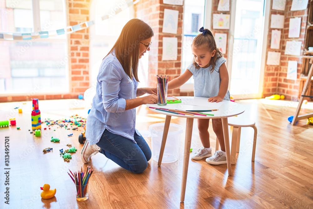 Caucasian girl kid playing and learning at playschool with female teacher. Mother and daughter at playroom around toys drawing with pencil colors Caucasian girl kid playing and learning at playschool with female teacher. Mother and daughter at playroom around toys drawing with pencil colors